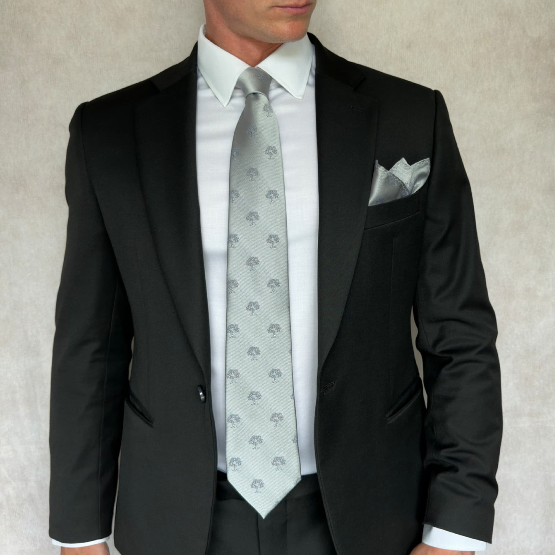 model wearing a black open suit and white shirt with grey and white tree embroidered tie and pocket square set